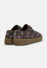 Zapatos de ante marrón sin cordones con bordado floral azul y una suela de goma texturizada en tono marrón claro. Presentan bordes suaves y un diseño sencillo.