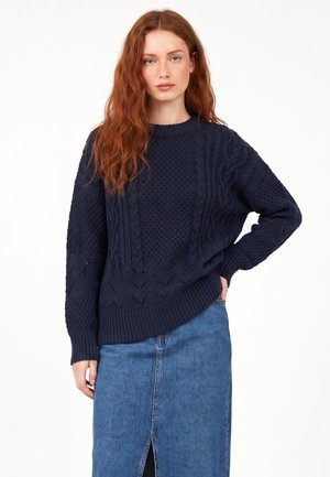 Giovane donna con lunghi capelli rossi che indossa un maglione lavorato a maglia blu navy con trama e una gonna di denim blu con uno spacco frontale.