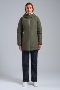 Donna in piedi, rivolta in avanti, indossa una giacca a zip verde oliva con cappuccio, jeans scuri e sneakers bianche, su uno sfondo neutro.