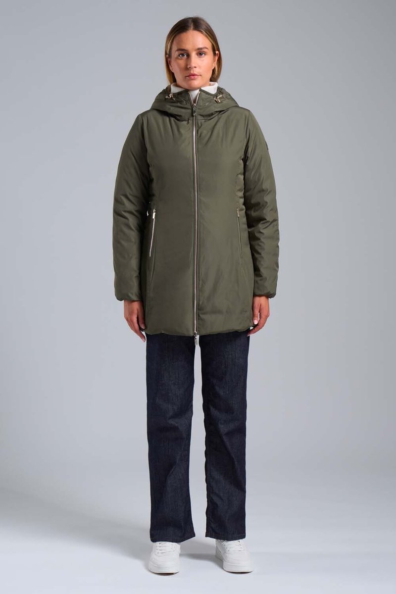 Donna in piedi, rivolta in avanti, indossa una giacca a zip verde oliva con cappuccio, jeans scuri e sneakers bianche, su uno sfondo neutro.
