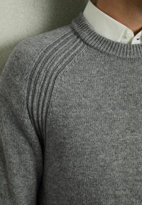 Grå stickad tröja med ribbad textur vid axelsömmen, med en klassisk rund halsringning och en slät finish.