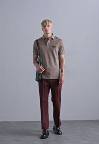 Brun polo skjorte med et logo, parret med kastanjefarvede bukser. Modellen bærer en grå skuldertaske og sorte sko. Simpelt design og rene linjer.