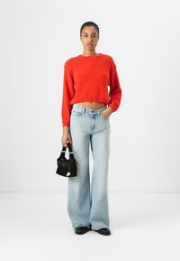 Pull court texturé rouge, jeans évasés bleu clair, et un petit sac à main noir. La modèle se tient devant un fond blanc uni.