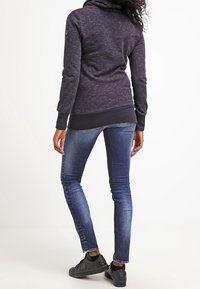 Žena ve tmavě šedém mikinovém svetru, úzkých modrých džínách a černých teniskách, stojící zády k divákovi na bílém pozadí.