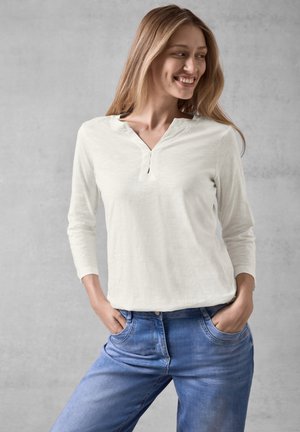 Světle krémový top s dlouhým rukávem, henley výstřihem a jedním knoflíkem, v kombinaci s džíny středně modré barvy. Hladká textura, volný střih.
