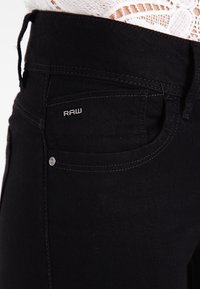 Musta farkkutakki, jossa on slim fit -leikkaus, huomaamaton ommel ja pieni hopeinen "RAW"-logo etulaukun yläpuolella.