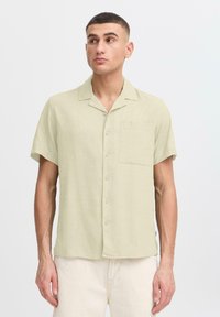 Chemise à manches courtes en lin vert clair avec une seule poche poitrine, fermeture à cinq boutons et coupe décontractée. Texture de tissu lisse.