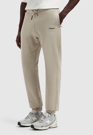 Man draagt beige joggerbroek met elastische boorden en een trekkoord in de taille, gecombineerd met grijze New Balance sneakers tegen een effen grijze achtergrond.