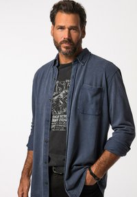 Camicia di jeans blu con bottoni sopra una maglietta nera con grafica, caratterizzata da una tasca sul petto, maniche lunghe e una vestibilità casual. Indossata con pantaloni scuri.
