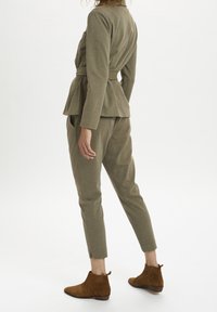 Olivgrüner, taillierter zweiteiliger Anzug mit einer Peplum-Jacke und schmal geschnittenen Hosen. Enthält braune Wildleder-Ankle Boots mit gerippten Details.