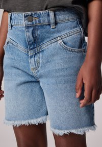 Jeans kratke hlače v svetlo modri barvi z raztrgano obrobo, obliko s petimi žepi, kontrastnimi šivi in gumbom za zapiranje. Opazne podrobnosti vključujejo kovinski gumb.