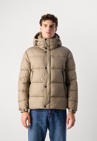 Doudoune beige avec capuche, présentant un design matelassé vertical, deux poches zippées latérales et des fermetures à boutons. Portée sur un jean en denim bleu.