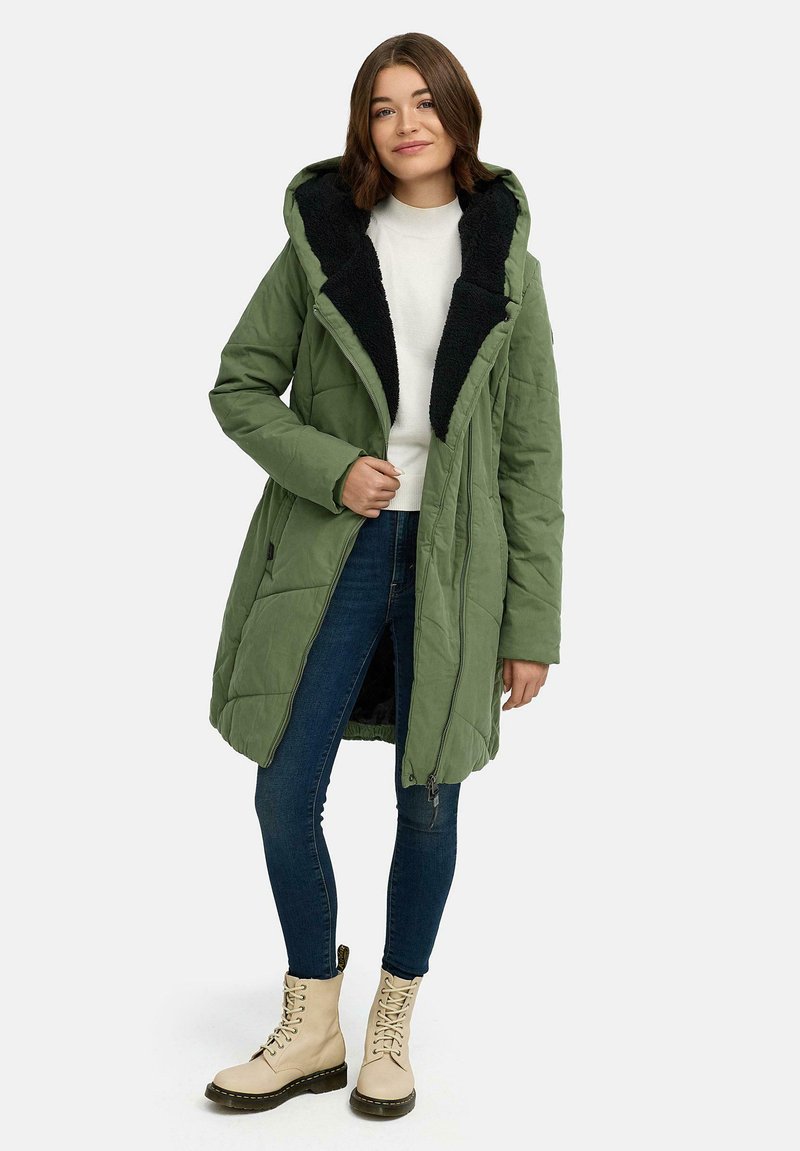 Grønn lang boblejakke med glidelås, vattert design, svart hette fôret med fleece og sidelommer. Brukt over en hvit genser og blå jeans.