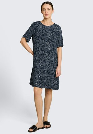 Frau steht, trägt ein dunkelblaues kurzärmliges Kleid mit kleinem weißem und gelbem Blumenmuster und schwarze Slide-Sandalen.