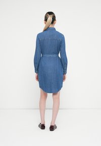 Kvinne med blond hestehår som har på seg en belted denim skjortekjole og brune skinnsko, står vendt mot en ensfarget hvit vegg.