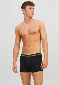 Schwarze Boxershorts mit einem gelben elastischen Bund, auf dem "JACK & JONES" steht. Der Stoff wirkt glatt und dehnbar. Das Modell posiert ohne Shirt.