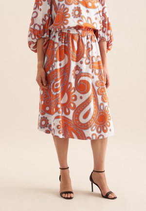 Jupe à motif floral en blanc, orange et bleu, dotée d'une taille élastique et d'un design fluide. Une paire de sandales à talons noires avec lanières est visible.