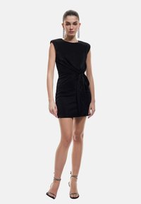 Robe mini noire ajustée en tissu texturé et scintillant ; elle possède des épaules larges et un détail en nœud torsadé à la taille.