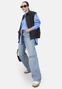Giacca nera senza maniche sopra una camicia azzurro chiaro, abbinata a jeans azzurri a gamba larga e sneakers bianche con suola beige. Occhiali da sole neri.
