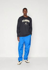 Juoda ilgomis rankovėmis marškinėliai su užrašu „MILWAUKEE“ ir logotipu, derinami su laisvomis mėlynomis kelnėmis ir neutralaus tono sportbačiais. Paprastas dizainas, laisvas siluetas.