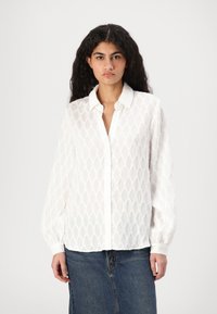 Camisa blanca de manga larga con un patrón de diamantes texturizado, cierre frontal de botones y un cuello clásico. Llevada con una falda de mezclilla.