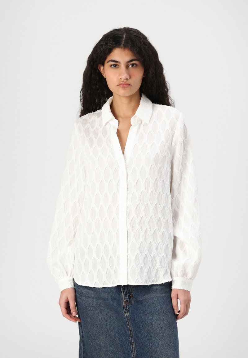 Camisa blanca de manga larga con un patrón de diamantes texturizado, cierre frontal de botones y un cuello clásico. Llevada con una falda de mezclilla.