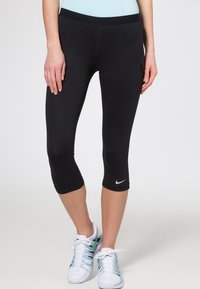 Schwarze Capri-Leggings mit glatter Textur, ausgestattet mit Seitentaschen und einem kleinen weißen Nike-Logo nahe dem Saum. Getragen mit weißen Sportschuhen.