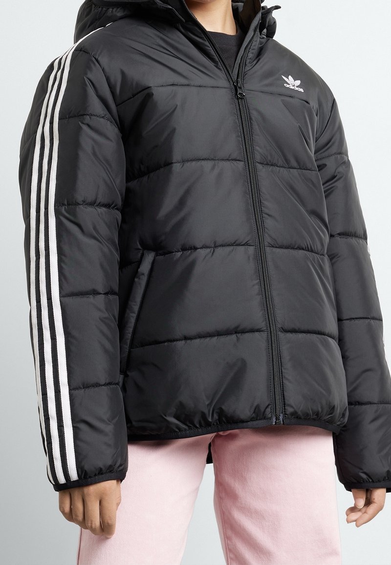 Person iført sort Adidas-puddet jakke med hvide striber på ærmerne og lyserøde bukser, der står foran en ensfarvet lys baggrund.