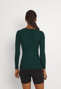Donna con i capelli intrecciati che indossa orecchini a cerchio dorati, un maglione a maniche lunghe a coste di colore verde scuro e pantaloni corti neri, voltata di spalle su uno sfondo bianco.