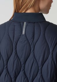 Giacca imbottita blu marina con collo alto, caratterizzata da un motivo a onde e un'etichetta grigia del marchio sul colletto. Tessuto morbido e liscio.