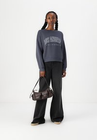 Felpa grigia oversized con stampa bianca, jeans neri a gamba larga e una borsa a spalla corta marrone con tasche, su uno sfondo semplice.