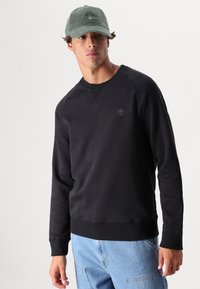 Felpa nera con collo rotondo e polsini a costine, caratterizzata da un piccolo logo sul petto. Abbinata a pantaloni in denim blu chiaro e un cappellino verde.