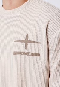 Primo piano di un maglione beige a coste con logo PXG marrone ricamato sul petto, che mostra parte della spalla e del collo.