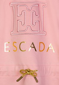 Tessuto rosa con logo ricamato "E" e la parola "ESCADA" in oro. Include un cordino con un fiocco decorativo dorato e punte metalliche.
