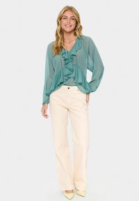 Teal, ruffled blouse with long sleeves paired with cream-colored, wide-leg jeans and light yellow heels. Smooth fabric, casual fit.