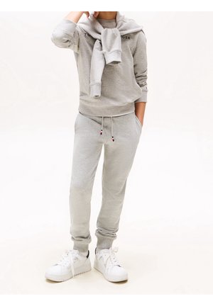 Person trägt hellgraue Sweatpants und Sweatshirt mit einem zweiten Sweatshirt, das über die Schultern gelegt ist, kombiniert mit weißen Sneakern.