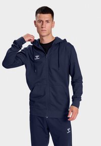 Navy zip-up hoodie with adjustable drawstring, front pockets, and logo detail on chest, paired with matching pants. Smooth fabric texture.