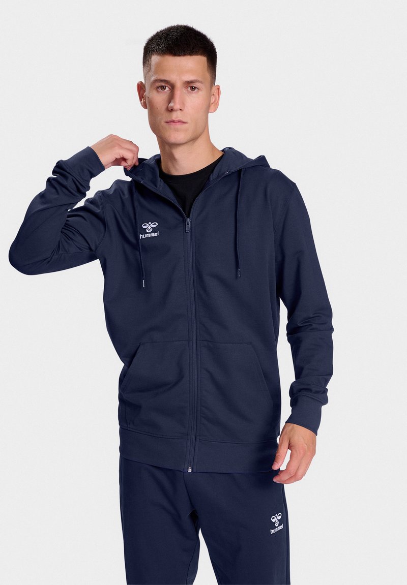 Navy zip-up hoodie with adjustable drawstring, front pockets, and logo detail on chest, paired with matching pants. Smooth fabric texture.