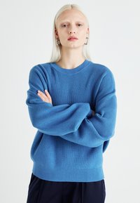 Kék kötött pulóver, kerek nyakkivágással, bordázott textúrával és lecsúszott vállakkal, hosszú ujakkal és laza fazonnal.