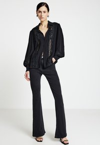 Schwarzes Hemd zum Knöpfen mit Spitzenbesatz, langen Ärmeln, lässigem Sitz; kombiniert mit schwarzen Schlagjeans und schwarzen offenen High Heels.