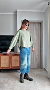 Χαλαρό πουλόβερ σε ελιά πράσινο με πέτρες ώμων, ανοιχτό μπλε φαρδιά τζιν και μαύρα μποτάκια μέχρι τον αστράγαλο. Λείο ύφασμα χωρίς ορατά σχέδια.