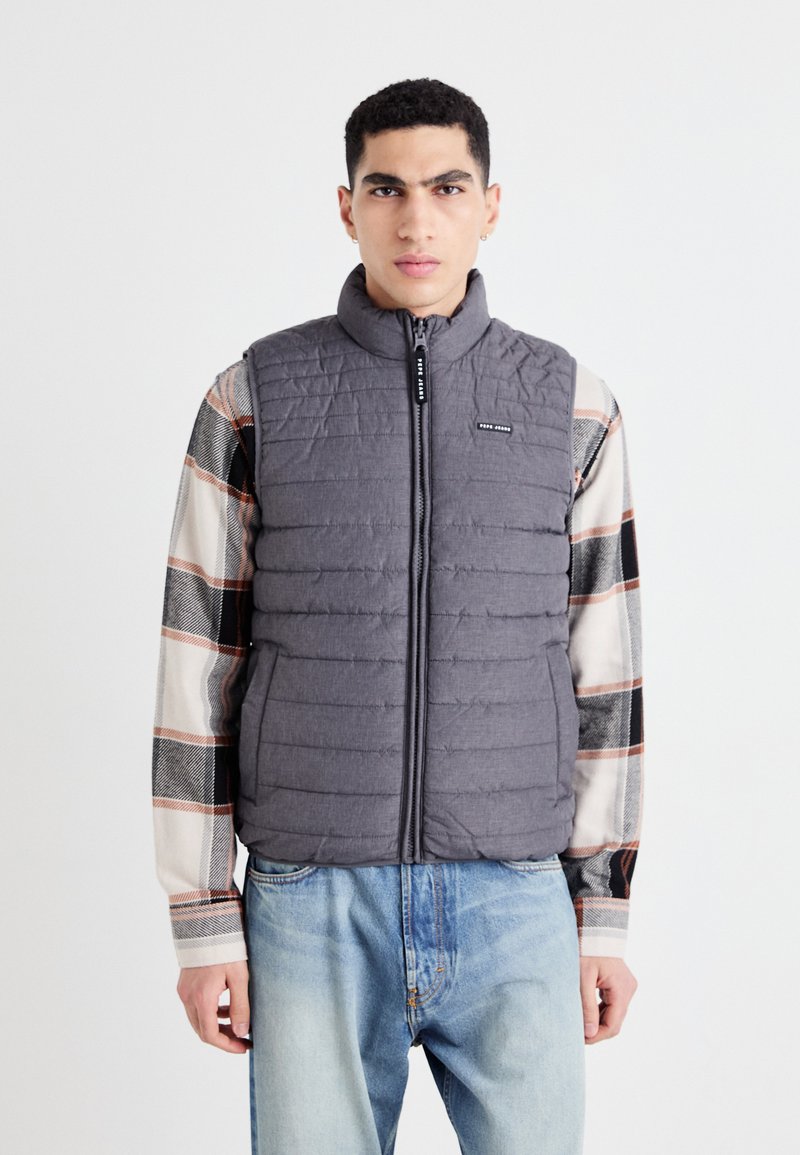 Chaleco acolchado gris con un cuello alto y cremallera frontal, llevado sobre una camisa de cuadros multicolores. Presenta un pequeño logo en el pecho.