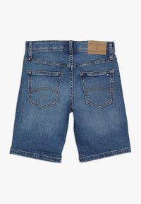 Denim-shortsit keskisinisessä värissä, suoralinjaisella leikkauksella, joissa on kaksi takataskua ja vaaleanruskea nahkavyötön tarra. Pehmeä pinta ja klassinen muotoilu.