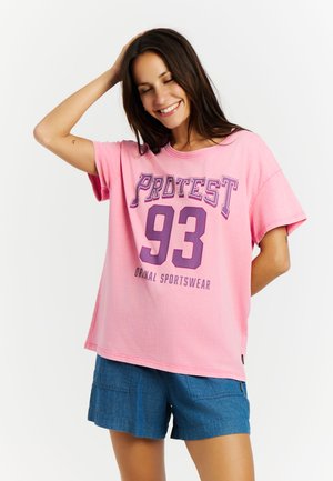 Glimlachende vrouw met lang donker haar, die een losse roze T-shirt draagt met "PROTEST 93" en een blauwe denimshort, poserend met één arm omhoog.