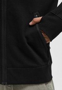 Giacca nera con zip a tutta lunghezza realizzata in un tessuto testurizzato, tasche laterali e polsini a costine. Presenta una zip liscia sul davanti per la chiusura.