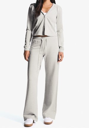 Mujer con sudadera con capucha de cremallera en gris claro y pantalones anchos a juego, con zapatillas blancas, sobre fondo blanco