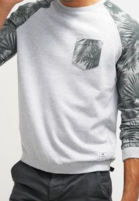 Uomo che indossa un felpa grigio chiaro con maniche a motivi di foglie di palma verde scuro e taschino coordinato, abbinato a pantaloni scuri.