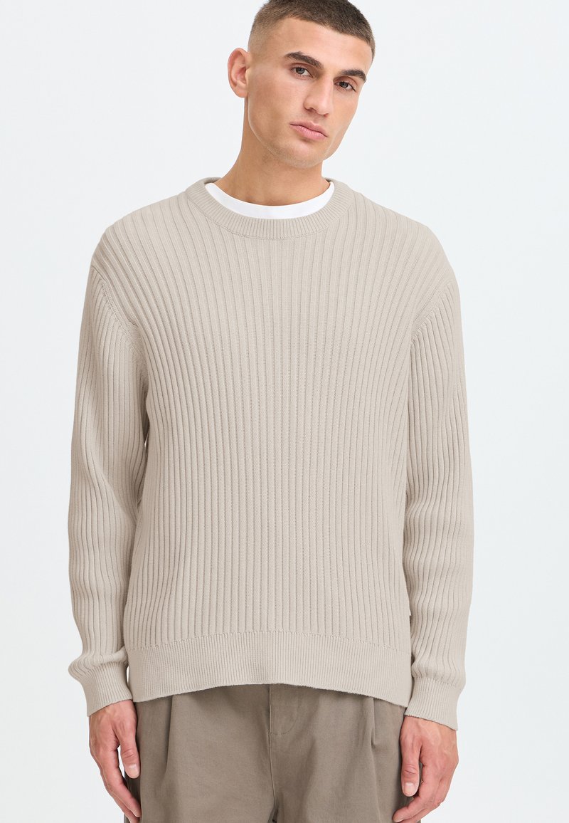 Jeune homme aux cheveux courts portant un pull côtelé beige par-dessus une chemise blanche et un pantalon marron clair, debout devant un fond blanc uni.