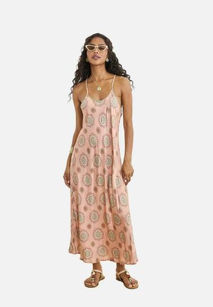 Mujer con cabello largo y rizado, vestida con un vestido maxi rosa con flores, gafas de sol beige de estilo ojo de gato, sandalias y joyería dorada sobre un fondo liso.