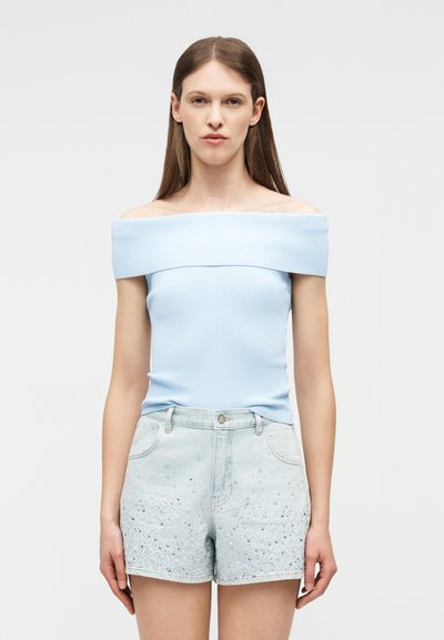 Γυναίκα που φορά γαλάζιο τοπ στράπλες και κοντό τζιν σορτς με μικρές στρας, στέκεται μπροστά σε απλό φόντο.